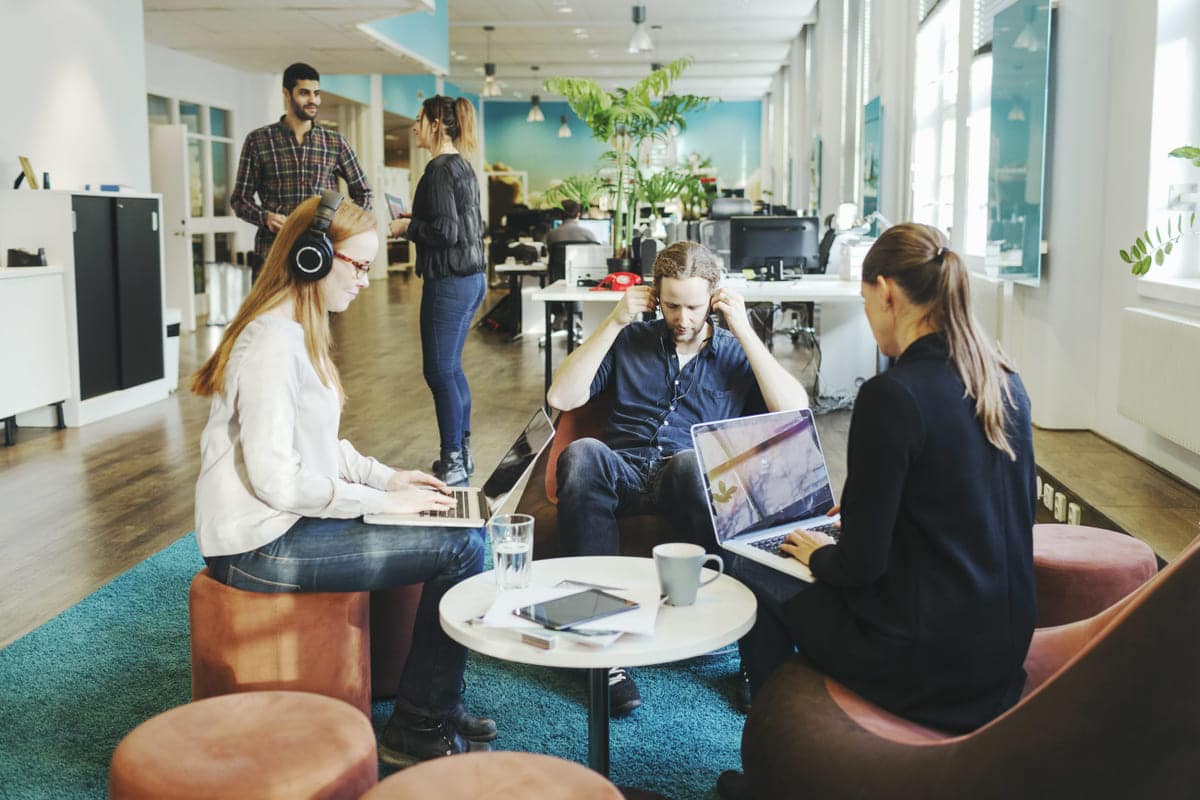 Tre personer sitter og jobber i åpent landskap, to kolleger står og prater like ved.