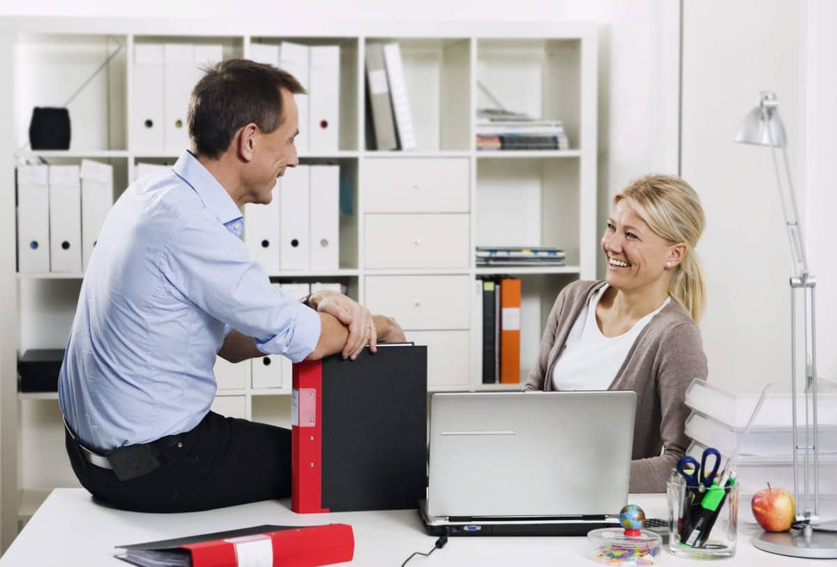 Hyggelig blid leder sitter på skrivebordet til en medarbeider. Begge smiler.