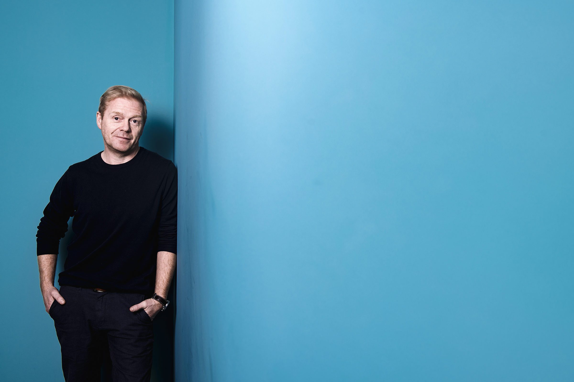 Blond mann, med hendene i bukselommene, står lent mot en blå vegg
