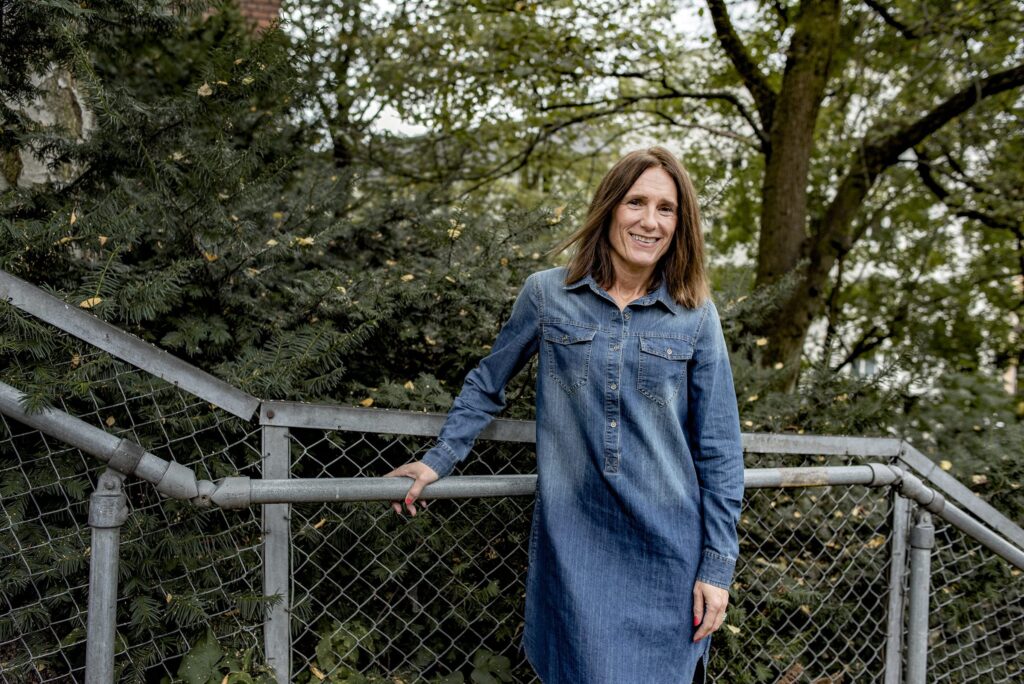 En kvinne med halvlangt brunt hår står smilende ved et gjerde. Hun har på seg en dongerikjole.
