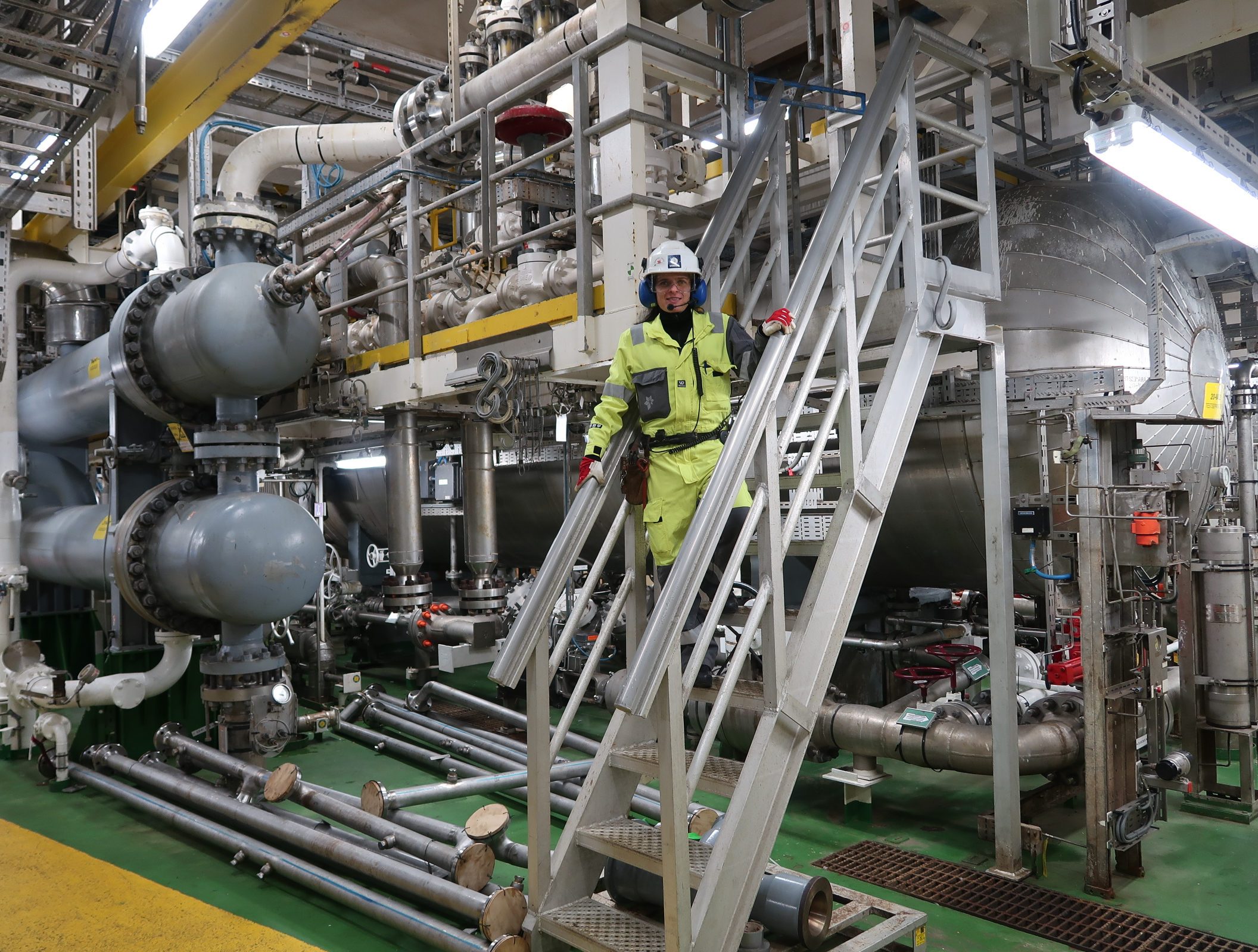 Kvinne i gule arbeidsklær og hjelm står i en trapp i et miljø preget av rør.