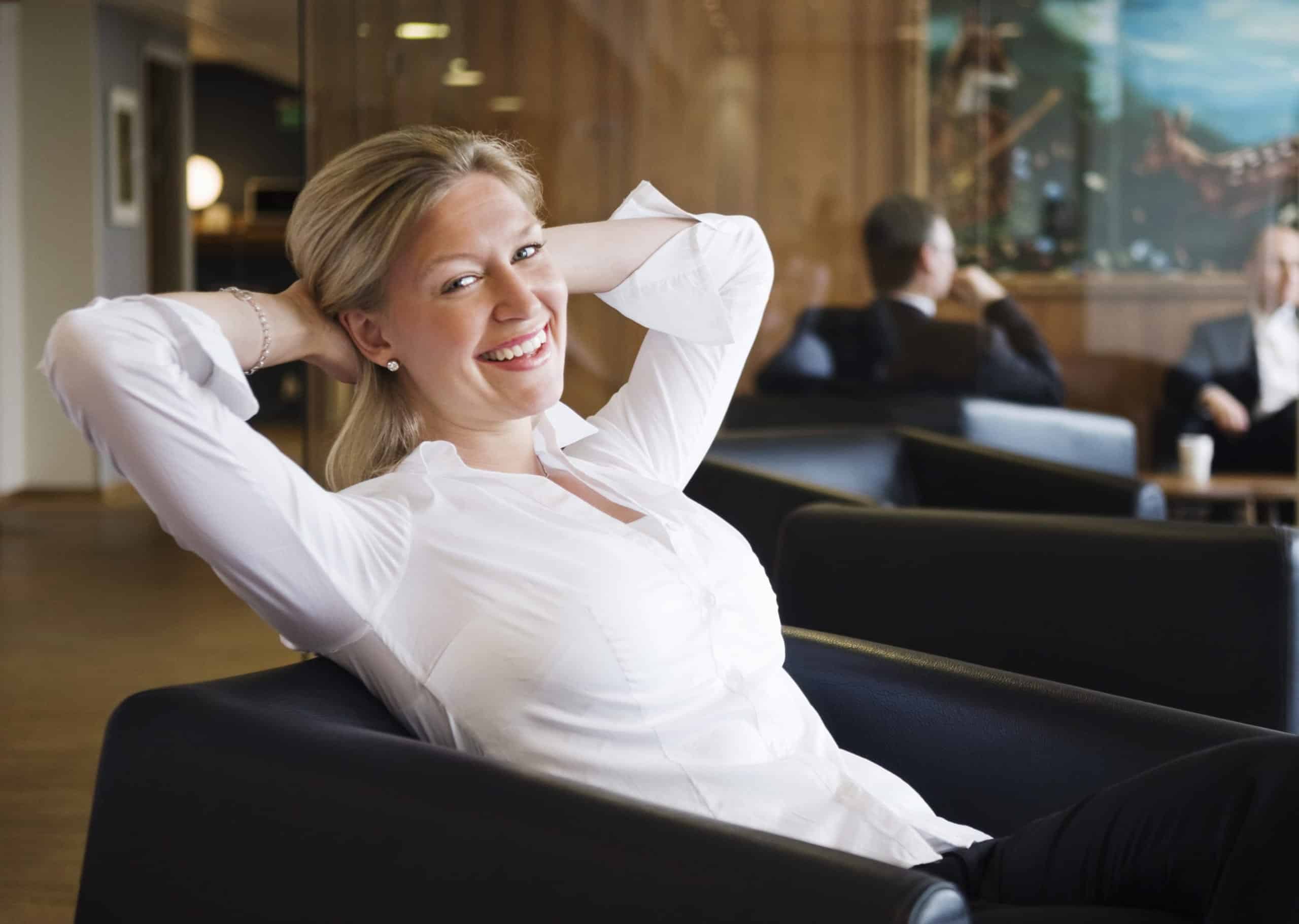 Kvinne i hvit skjorte sitter tilbakelent og ser fornøyd ut. I bakgrunnen anes et par menn.