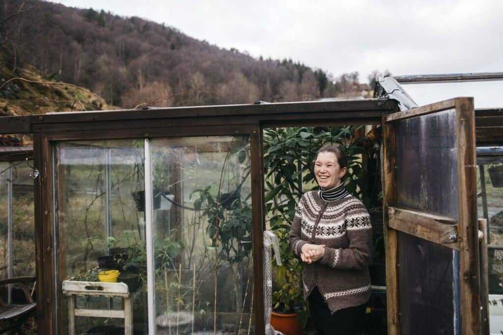 Ung kvinne i tradisjonell strikkekofte står i inngangen til et lite drivhus.