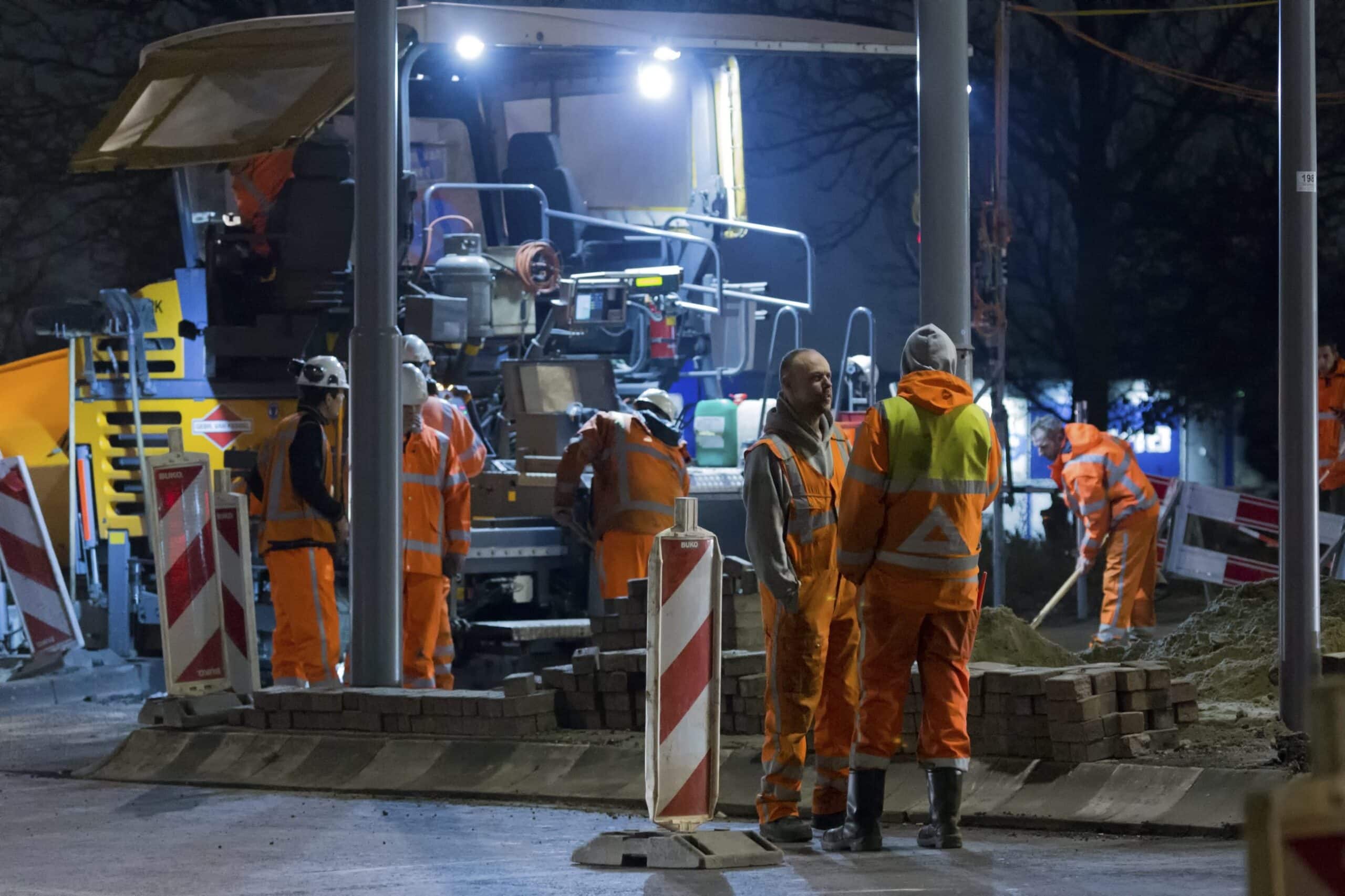En gruppe anleggsarbeidere i orange klær jobber i mørket. En maskin og noen skilt er også med i bildet.