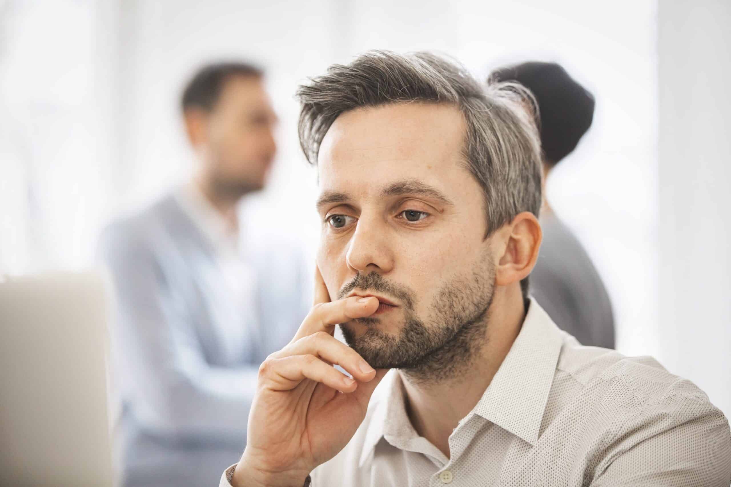 Mann på cirka 40 år, med grått hår og skjegg, sitter med en hånd foran munnen og ser tankefull ut. To andre anes i bakgrunnen.