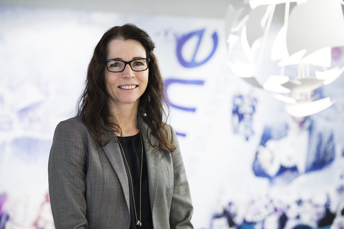 Suksess. Kvinne med langt brunt hår, briller og grå blazer står foran en vegg.