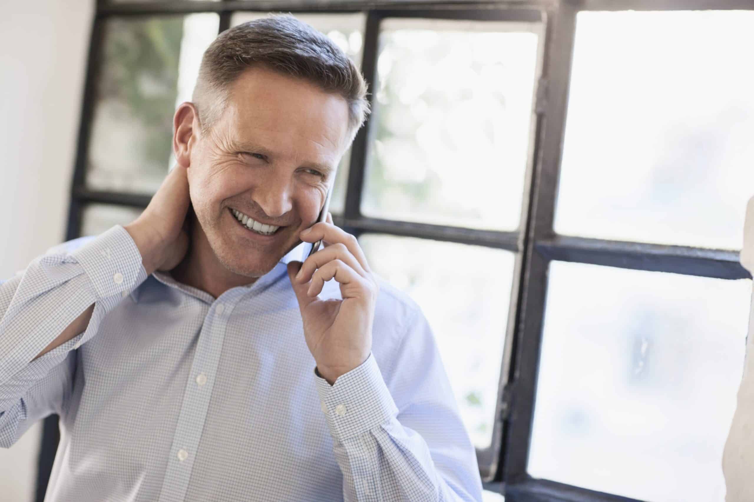 Blid mann med lys skjorte står foran et vindu og prater i telefonen.