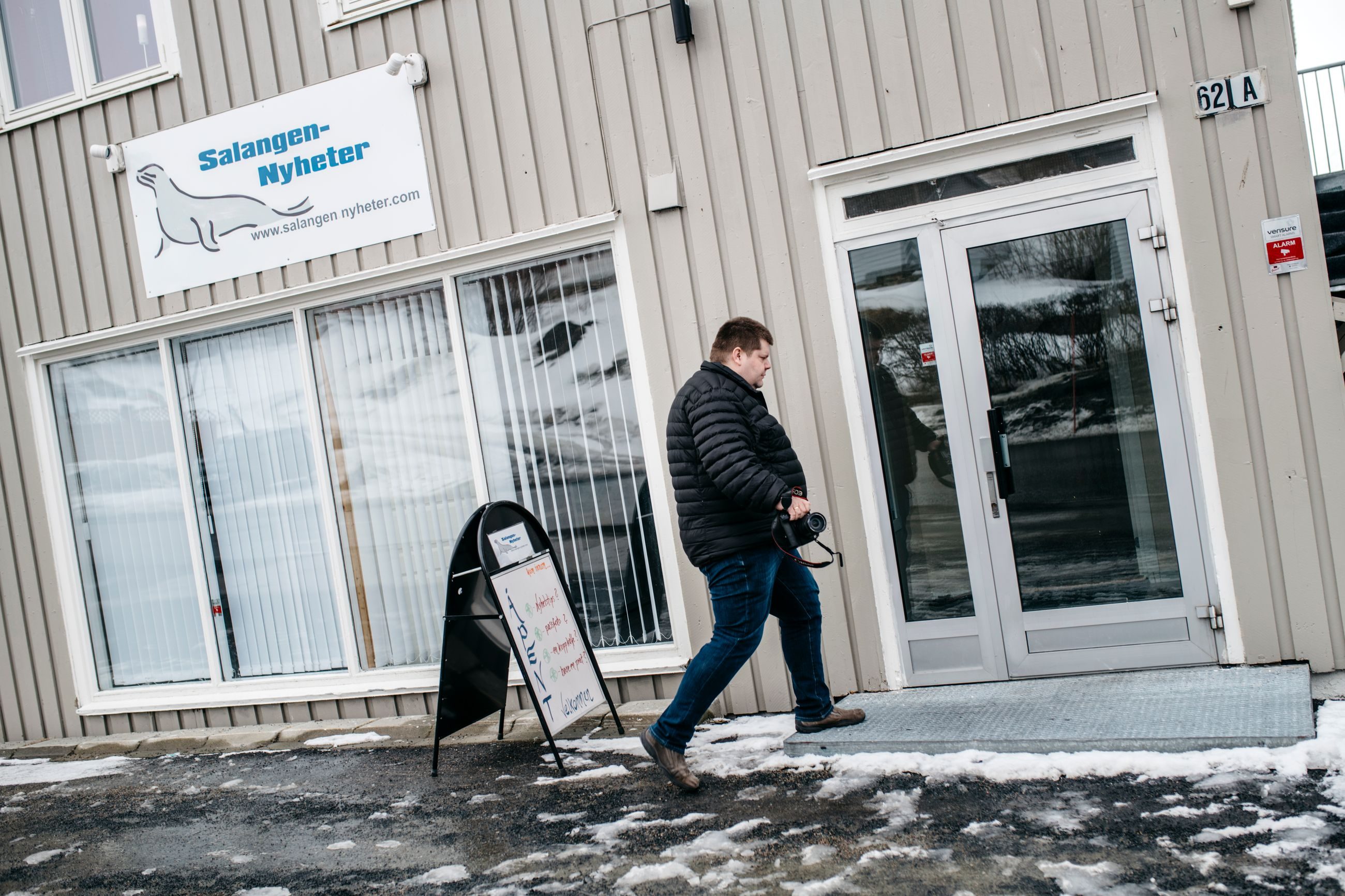 Mann i boblejakke på vei inn i et grått hus. På veggen står det Salangen Nyheter.
