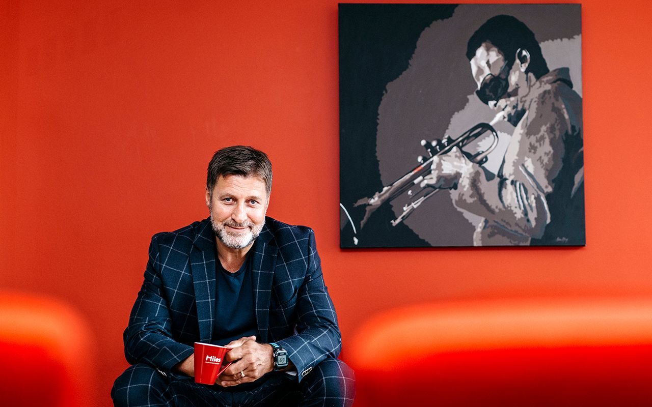 Verdier: Mann sitter i en sofa, og holder en rød kopp. Interiøret er rødt, og på veggen henger det et bilde av en mann som spiller trompet.