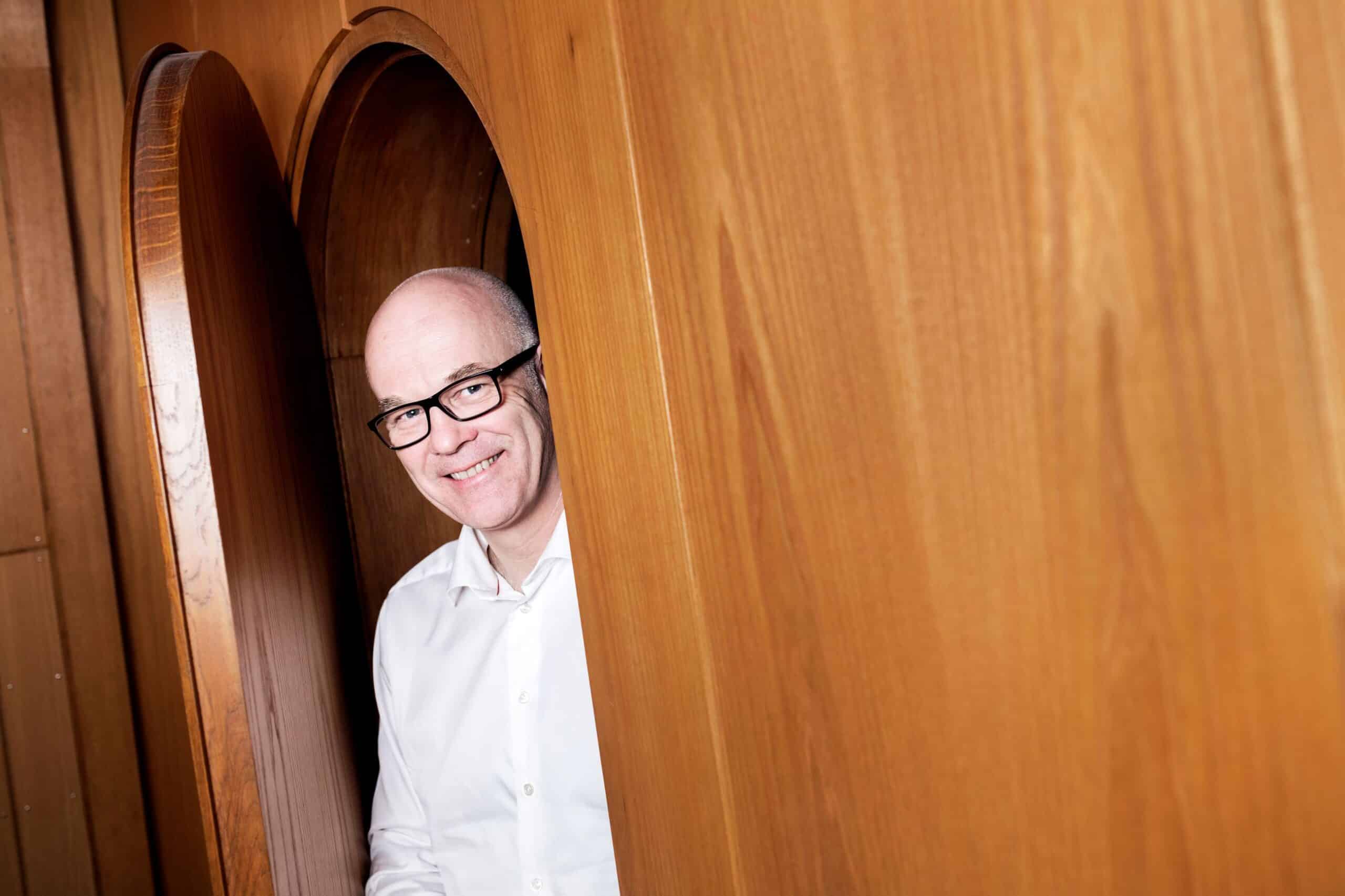 Nysgjerrig: Mann med briller og barbert hode titter ut gjennom en tredør.