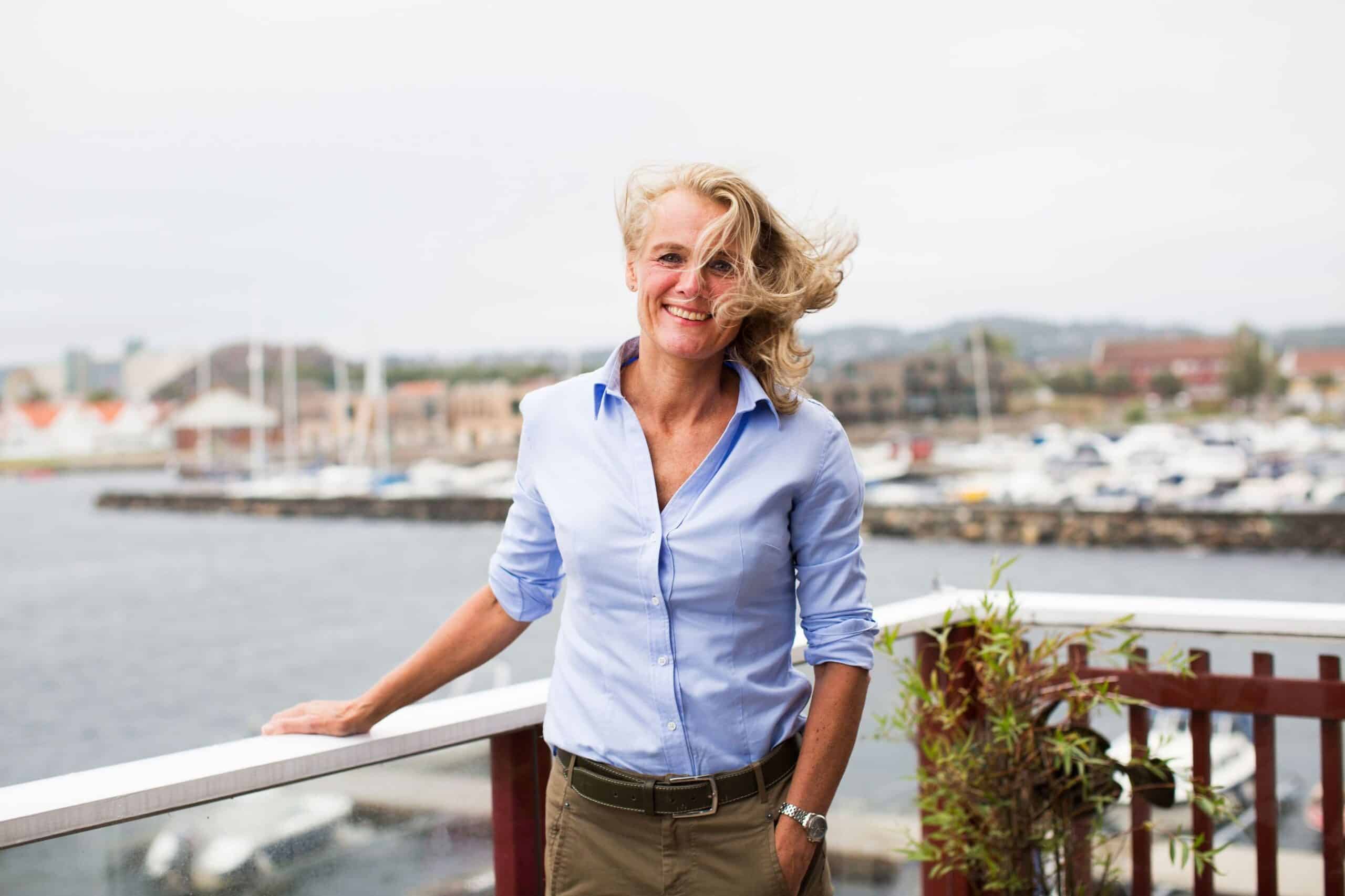 Endring: Blond kvinne står på en veranda med en småbåthavn i bakgrunnen.