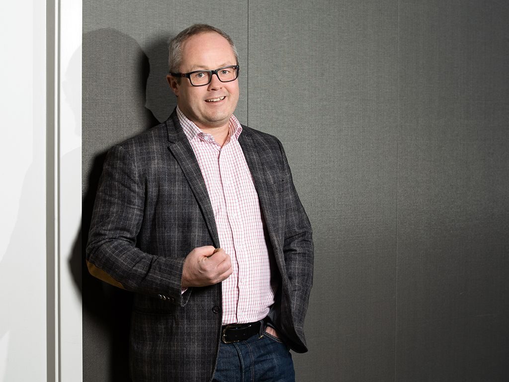 Sykefravær: Mann i grå jakke står foran en vegg.