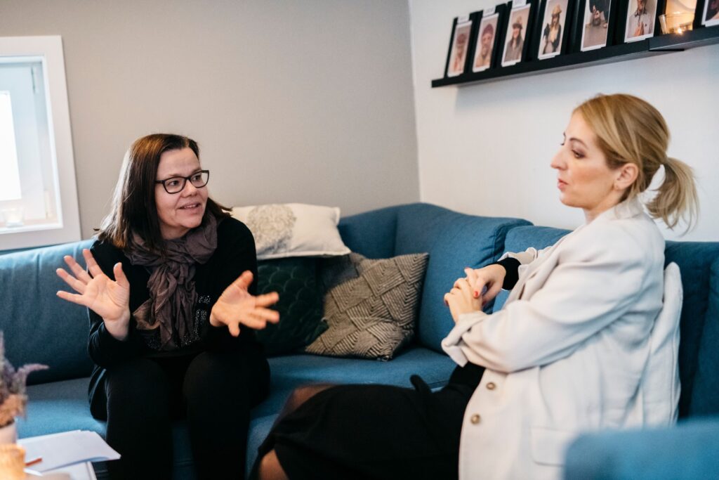 Sykefravær: To kvinner sitter i en hjørnesofa og snakker sammen.