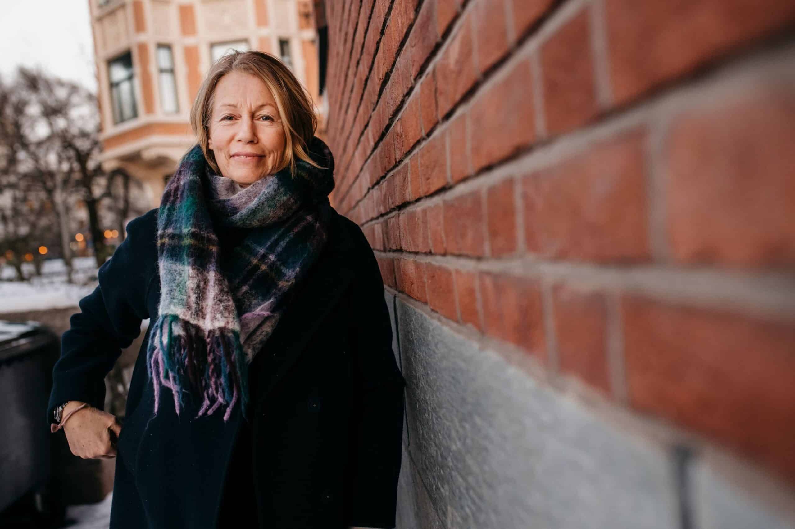 Verdier: Voksen kvinne står lent mot en mursteinsvegg.