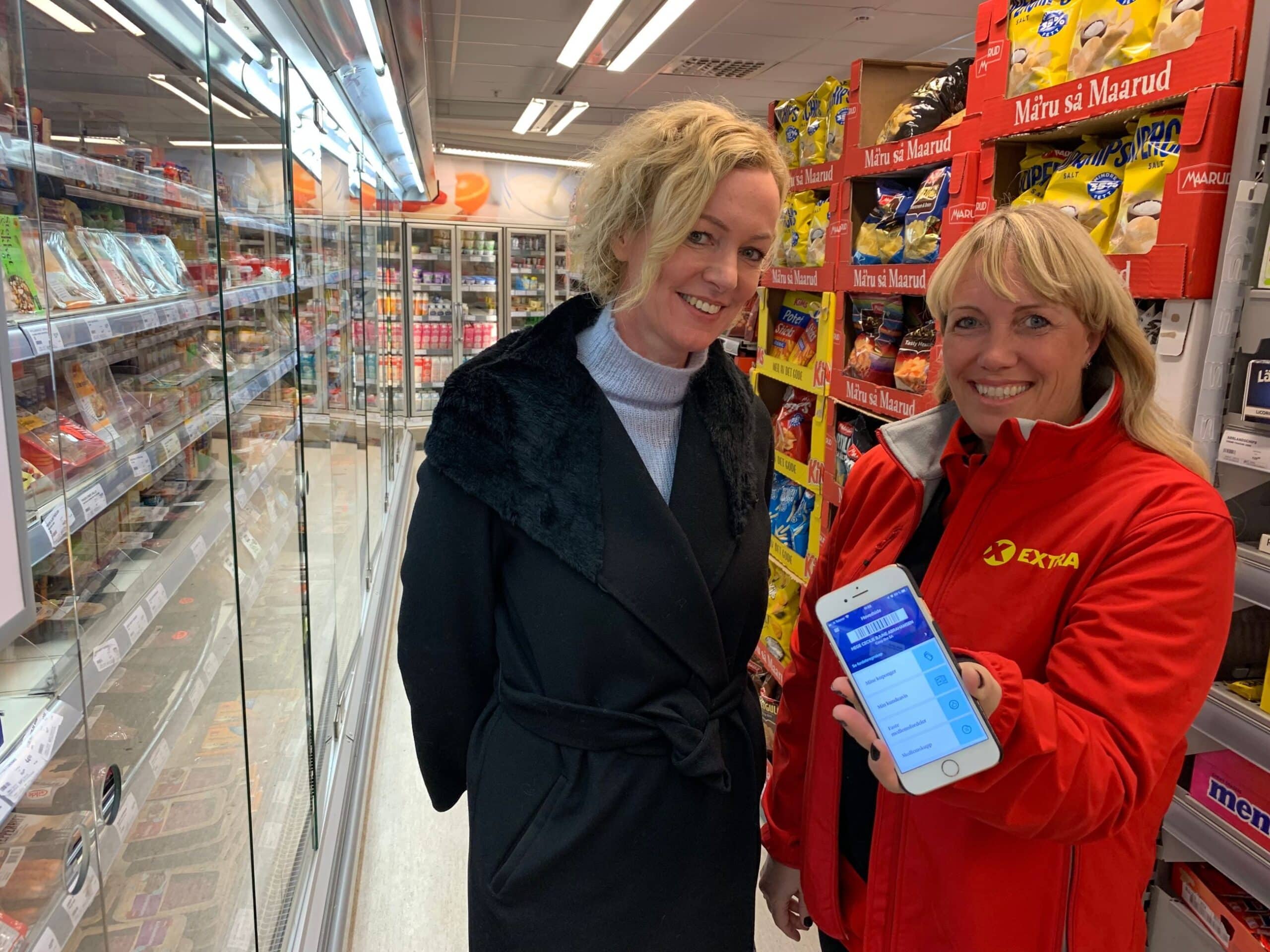 Digitalisering: To kvinner står i en matbutikk og holder fram en mobiltelefon.