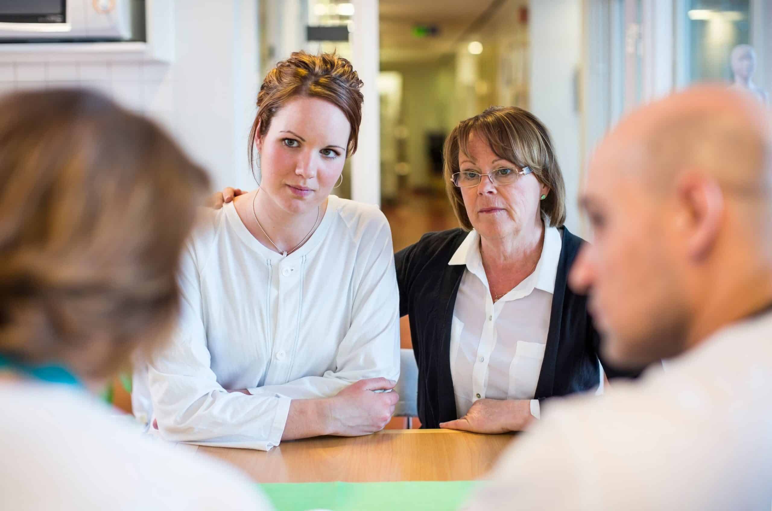 En ung kvinne og en godt voksen kvinne sitter sammen og snakker med helsepersonell.