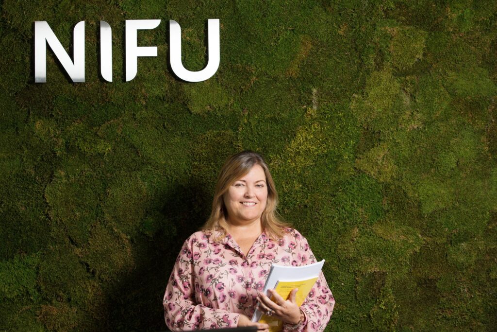 Seniorene: Blid kvinne står og smiler foran en mosevegg.