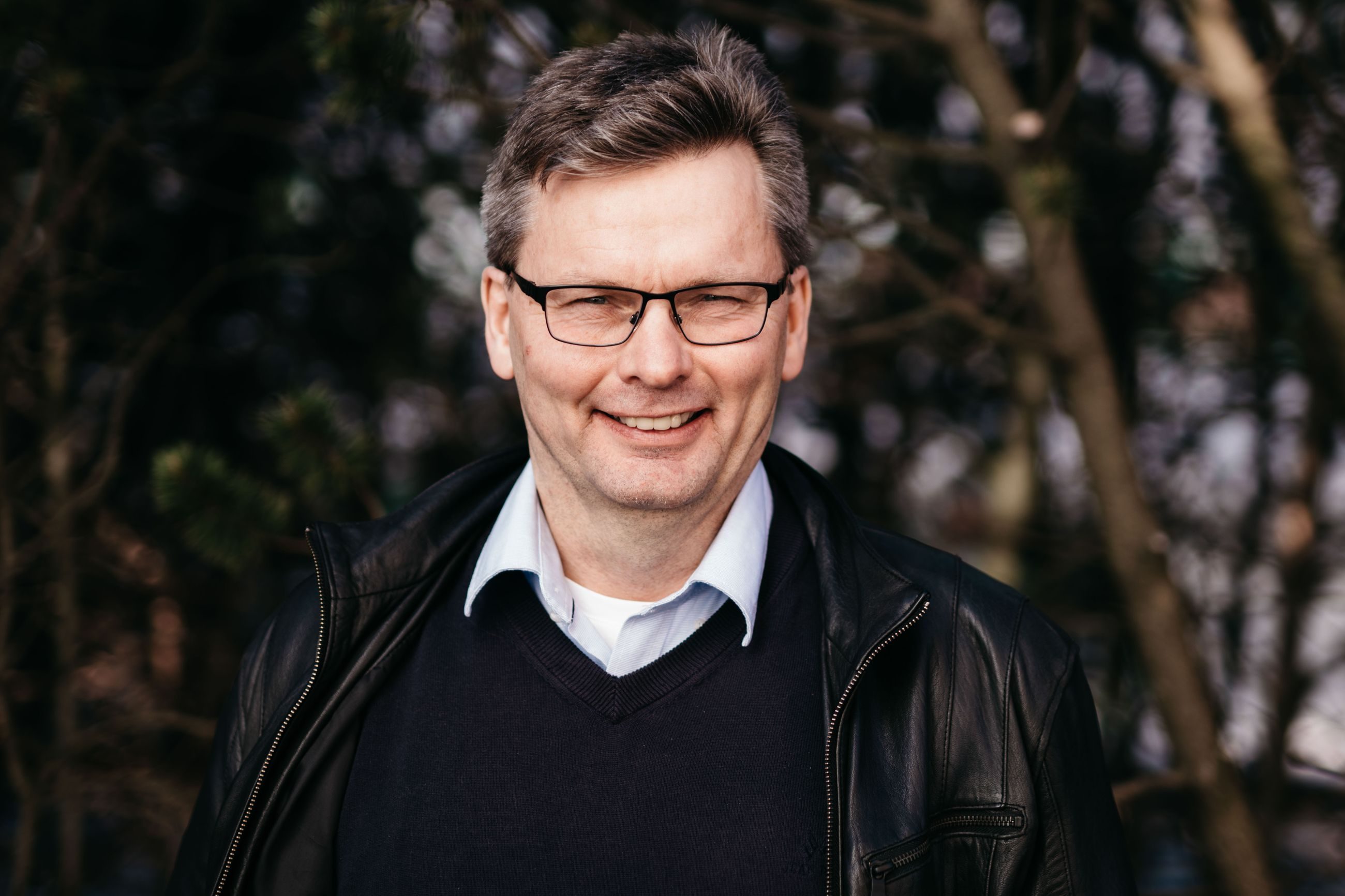 Ledere: Blid mann står utendørs foran en hekk.