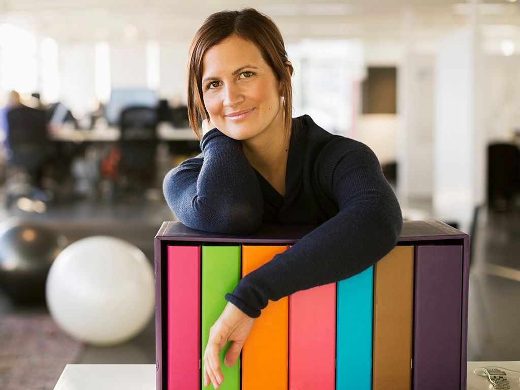 Ledere: Kvinne står lent over en hylle i et kontorlandskap, og smiler til fotografen.