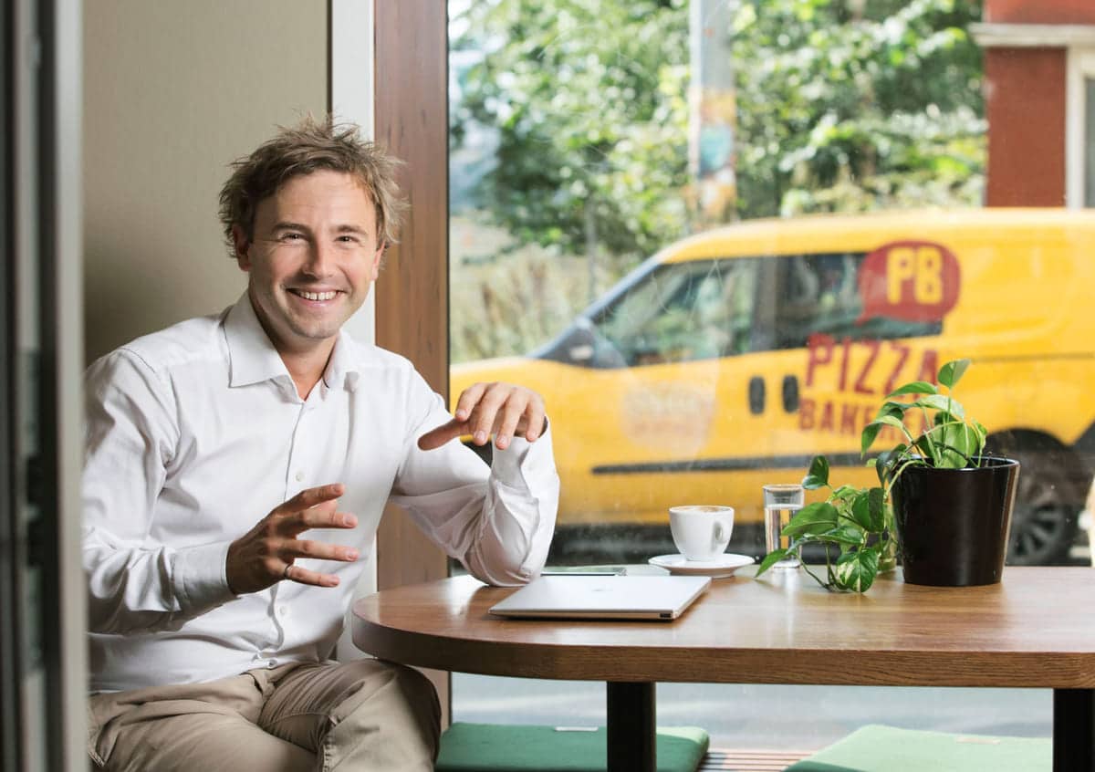 Kulturer: Blid mann sitter ved et bord, og utenfor kjører en gul pizzabil forbi.