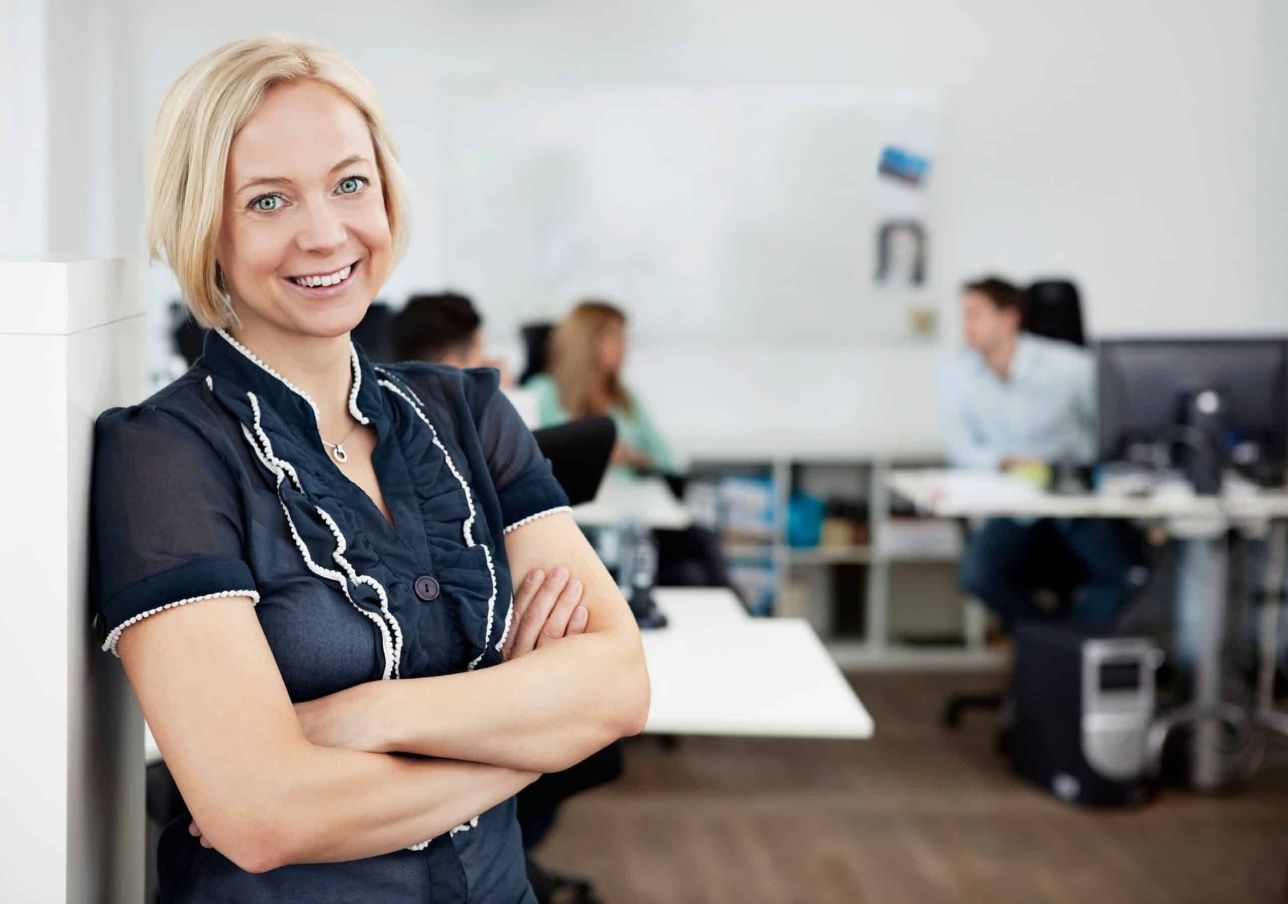 Blid, blond kvinne står i forkant og noen kolleger sitter i bakgrunnen.