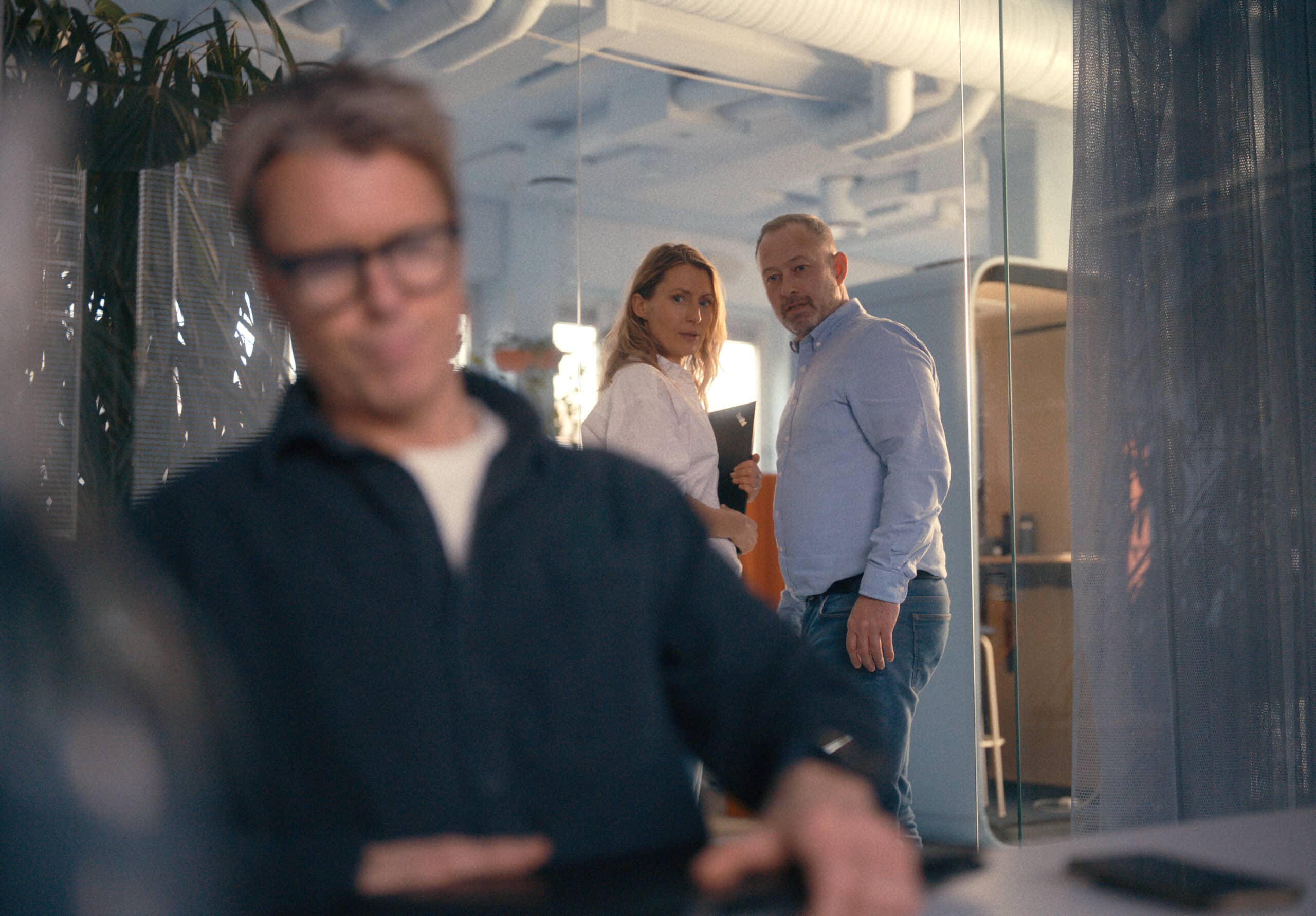 Alvorlig mann som sitter. To mennesker står på utsiden og skuler.