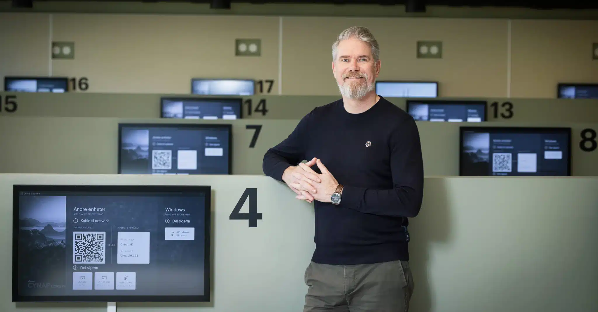 Eivind Brevik står foran en rekke pcskjermer og smiler til kamera