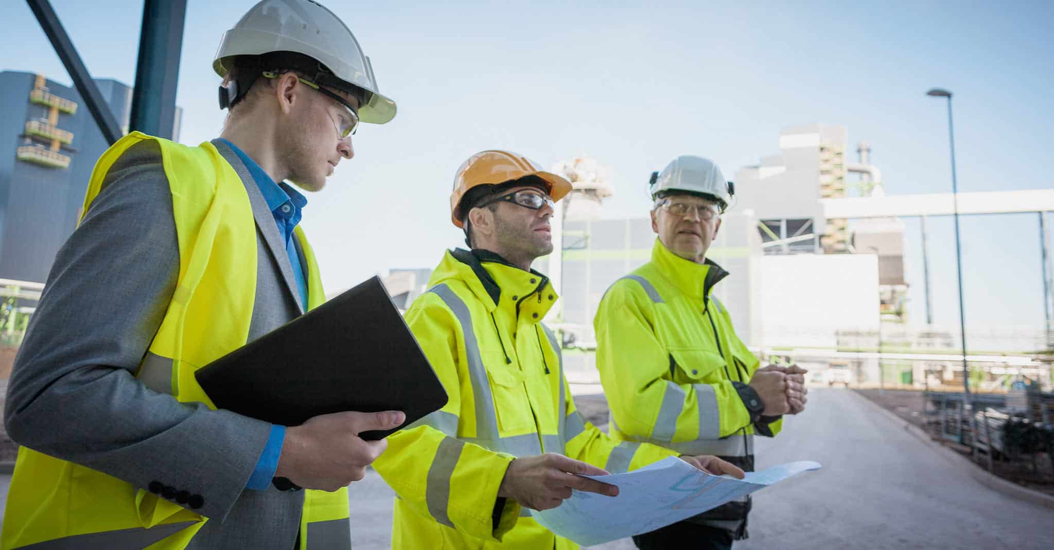 Tre menner står og titter på et prosjekt, ikledd hjelm og refleksvester