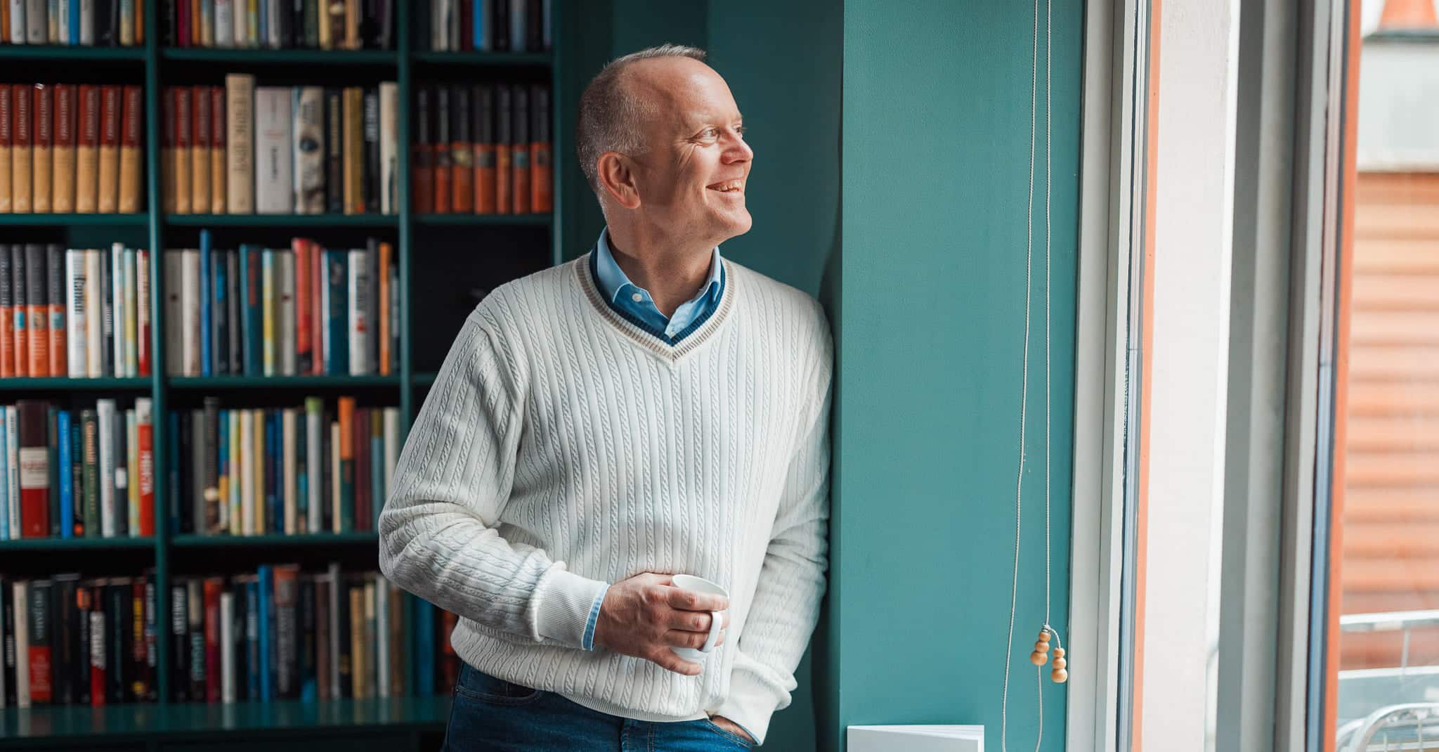 Stein Wesenberg titter ut av vindu med en kaffekopp i hånda og en bokhylle bak seg